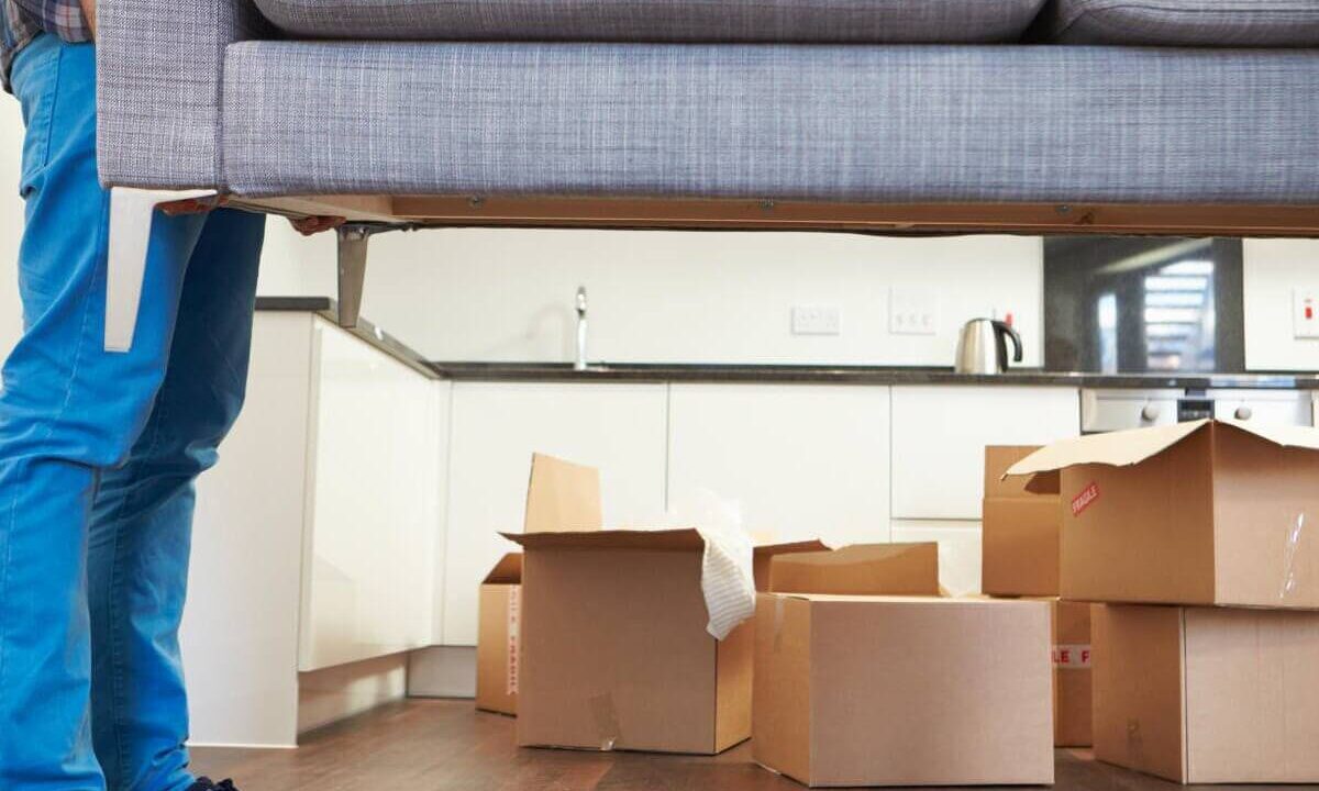 A photo showing a man lifting a sofa with some moving boxes in the background.