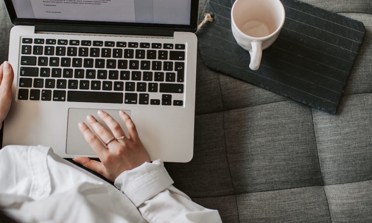 A photo showing a woman sitting on a sofa and using a laptop with a coffee next to her.