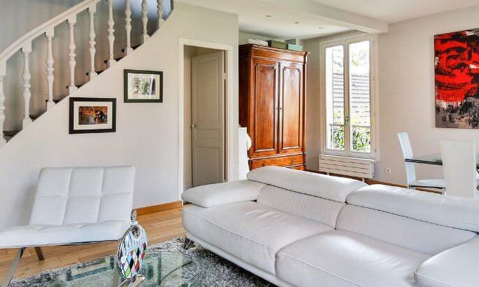 A photo showing a living room with a sofa, armchair, dining table and chairs and a large painting on the wall, with stairs on the left.