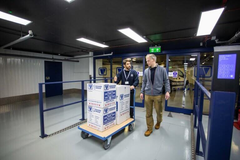 A photo showing a Vanguard employee pushing a trolley with Vanguard cardboard boxes on it for a customer moving into one of the self-storage units.