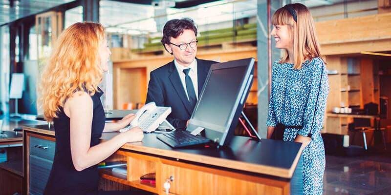 A photo showing two people, a man and a woman, talking to a receptionist.