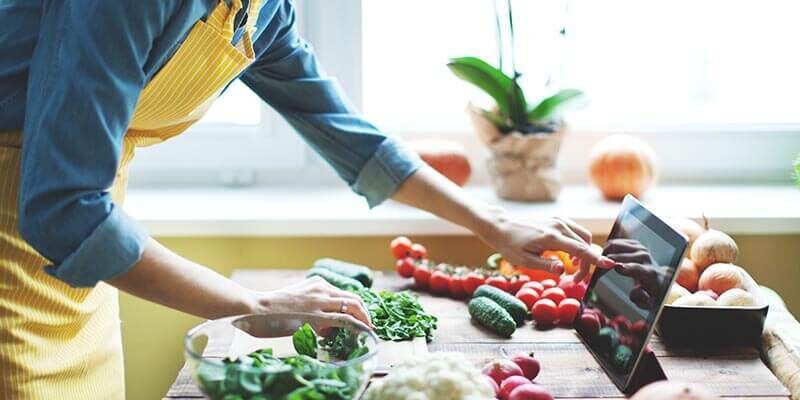 A photo showing a person wearing an apron, reading a recipe on a tablet with an array of vegetables on the kitchen counter in front of them.