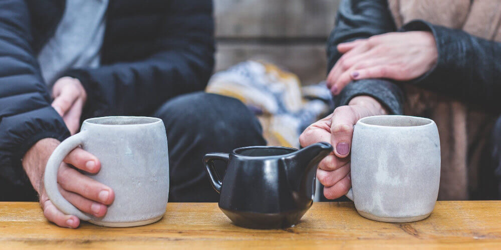 A photo of two people drinking tea together. The photo shows two people holding grey mugs with a black teapot in the middle.