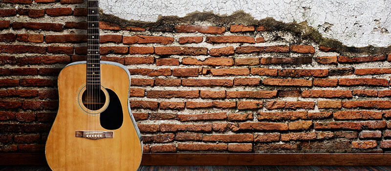 A photo showing an acoustic guitar resting in front of a brick wall.