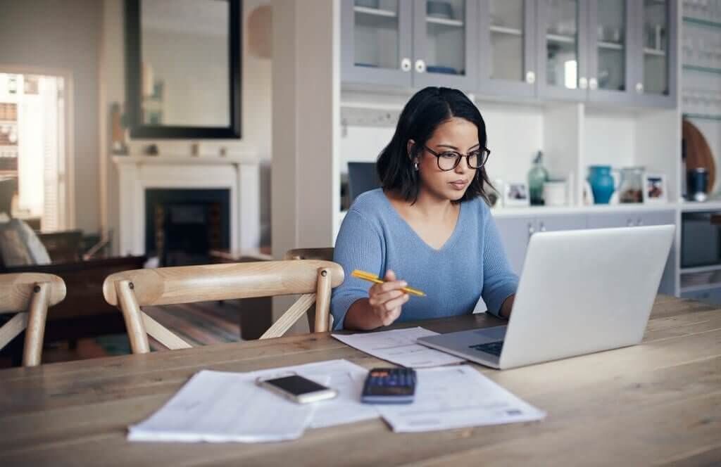 A photo showing a woman in front of a laptop, holding a pen with papers, a phone and a calculator next to her.