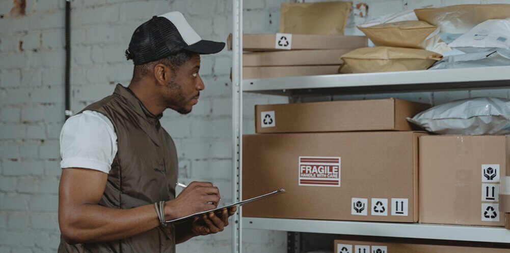 A photo showing a man holding a pen and paper and doing inventory management next to some shelves.
