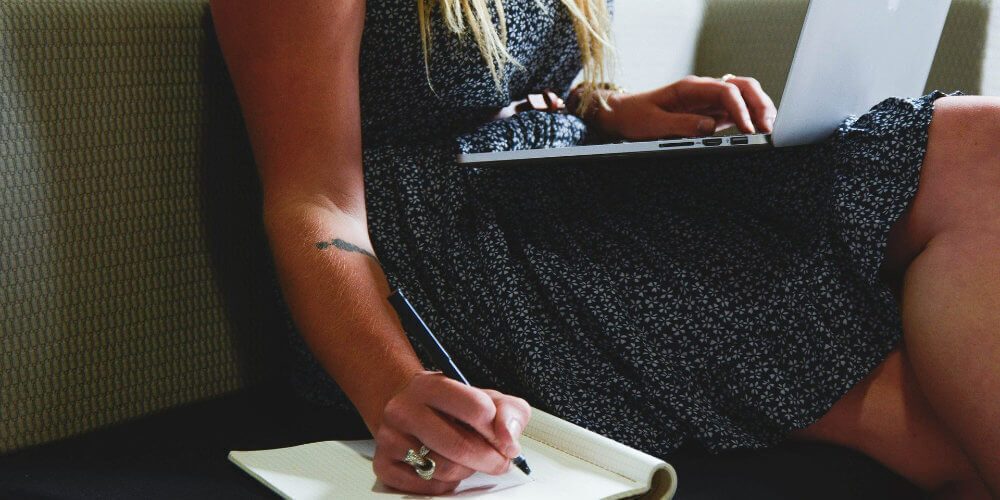 A photo showing a woman using her laptop and taking notes with a pen on a notebook.