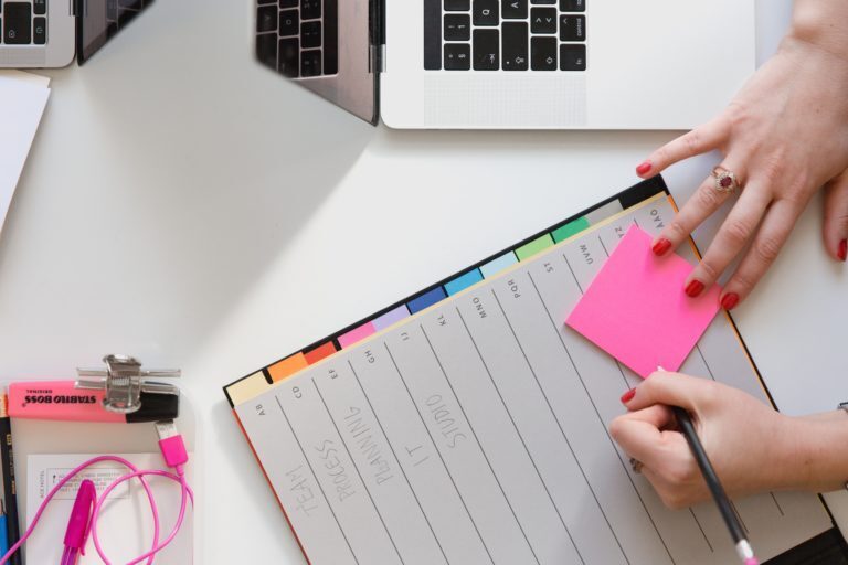 A photo showing a woman writing on a post-it note on top of an organiser book with a laptop next to her.