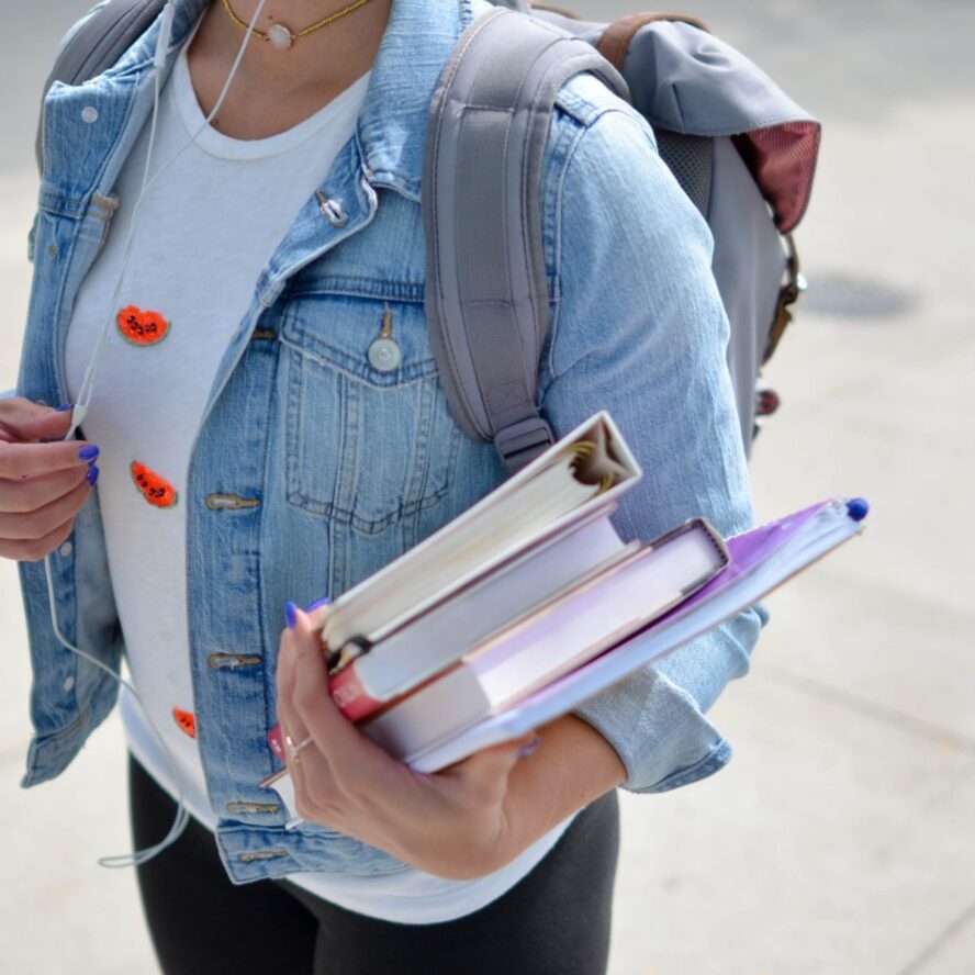 A photo showing a student wearing a backpack and holding a few books and binders in her arm.
