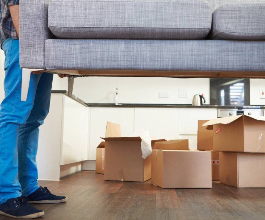 A photo showing a man lifting a sofa with some moving boxes in the background.