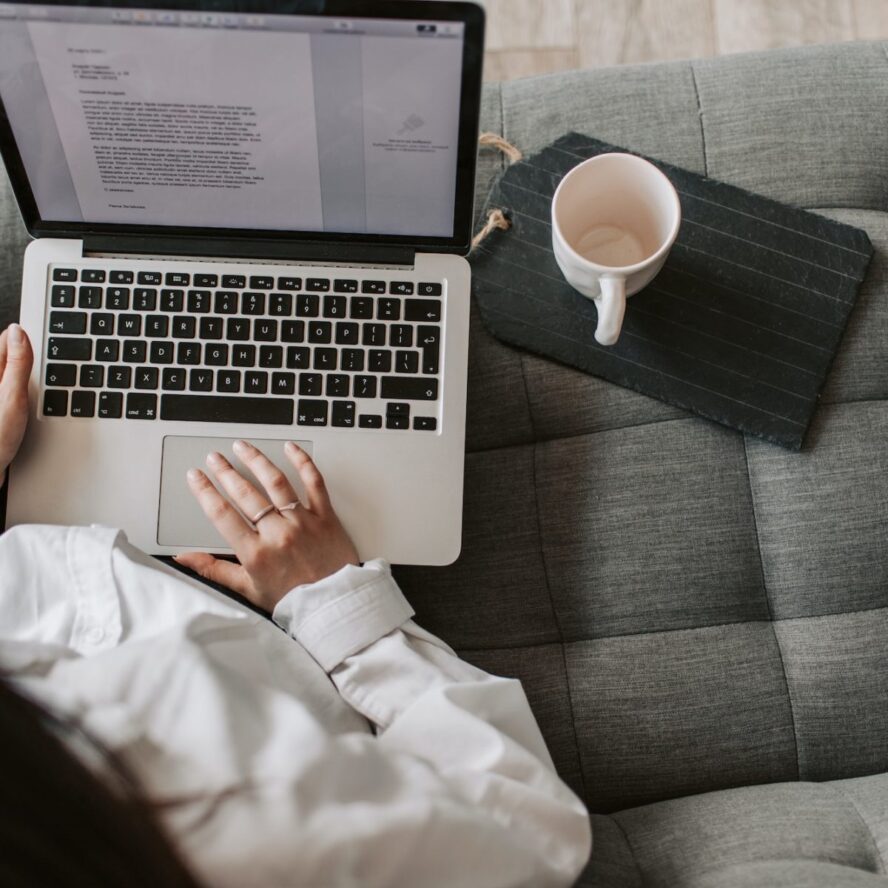 A photo showing a woman sitting on a sofa and using a laptop with a coffee next to her.