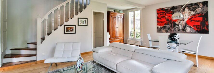 A photo showing a living room with a sofa, armchair, dining table and chairs and a large painting on the wall, with stairs on the left.
